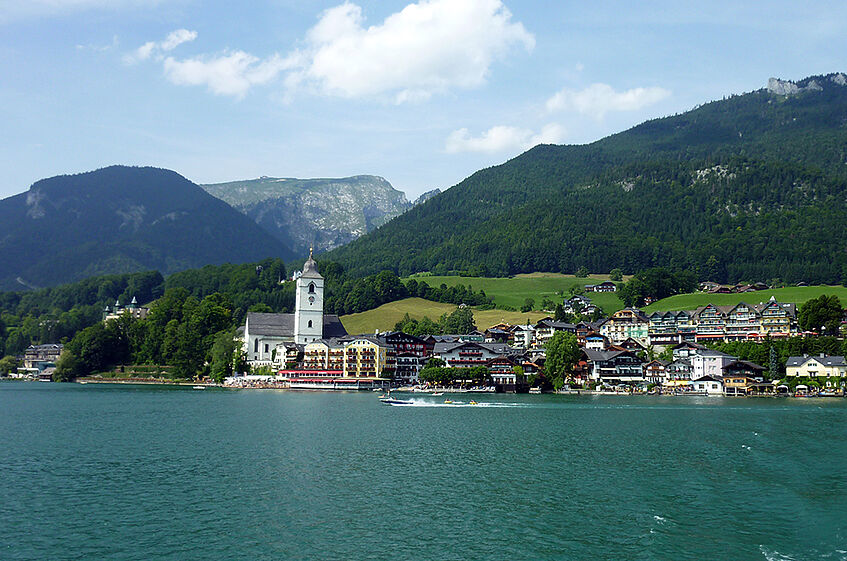 The village St. Wolfgang seen from the lake, with water in the foreground and a mountain range in the background.