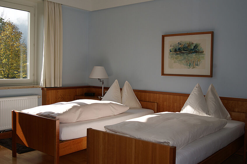 Spacious room with light blue walls, light curtains, and two wooden twin beds with white linnen.