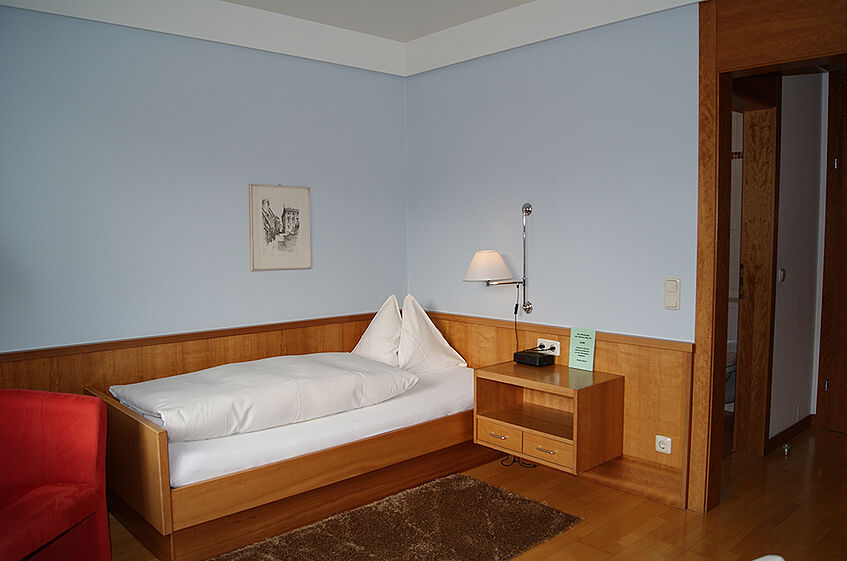 Spacious oom with light blue walls, a wooden single bed with white linnen, and a build-in night stand.