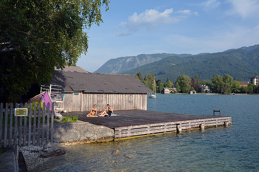 Boathouse with wooden deck at the shore of a lake with three people sitting in the sun.