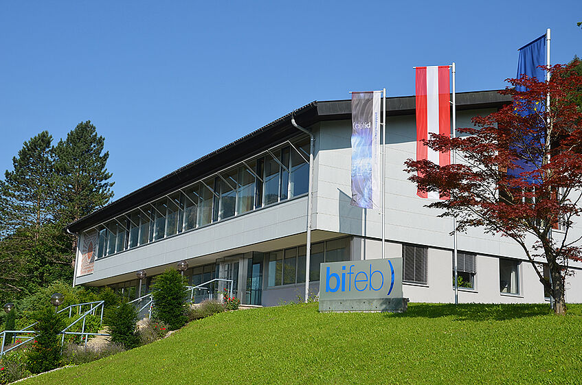 Two floored building on a slight grassy hill with flags of Austria and Europe on the side.