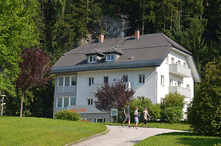 View of a White three story building, surrounded by a park area with meadows and trees and three people walking in front of the building.