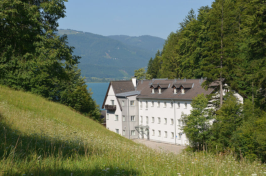 White four storey building in a park area surrounded by woods and a lake and mountains in the Background.