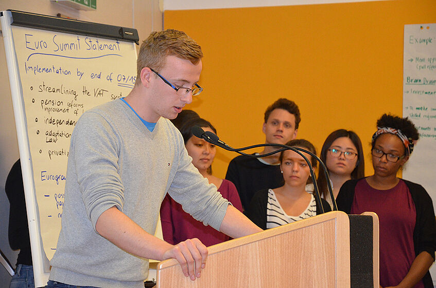 A student standing at a speaker's desk giving a presentation, with five students in the background.