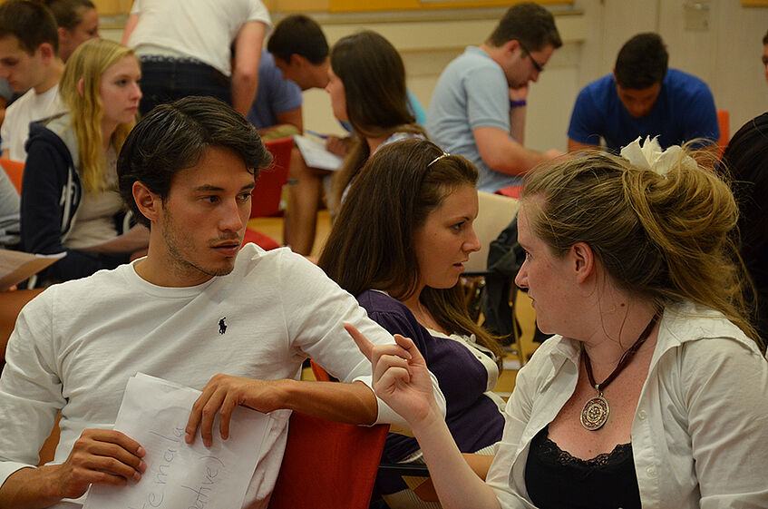 Two students involved in a discussion, in the background students working in groups.