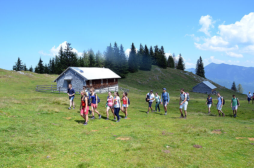 A group of young people walking accross a picturesque mountain pasture with an alpine cabin in the background.